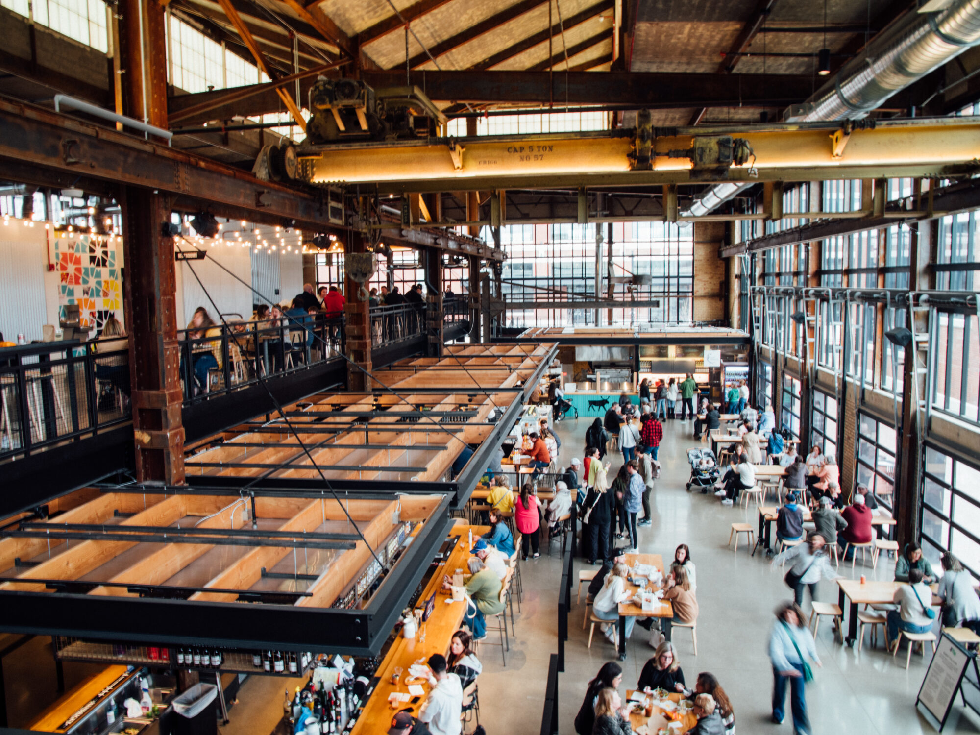 Industrial loft-style café or event space with exposed beams, large windows, string lights, and people seated at long communal tables above a bar area on the left.