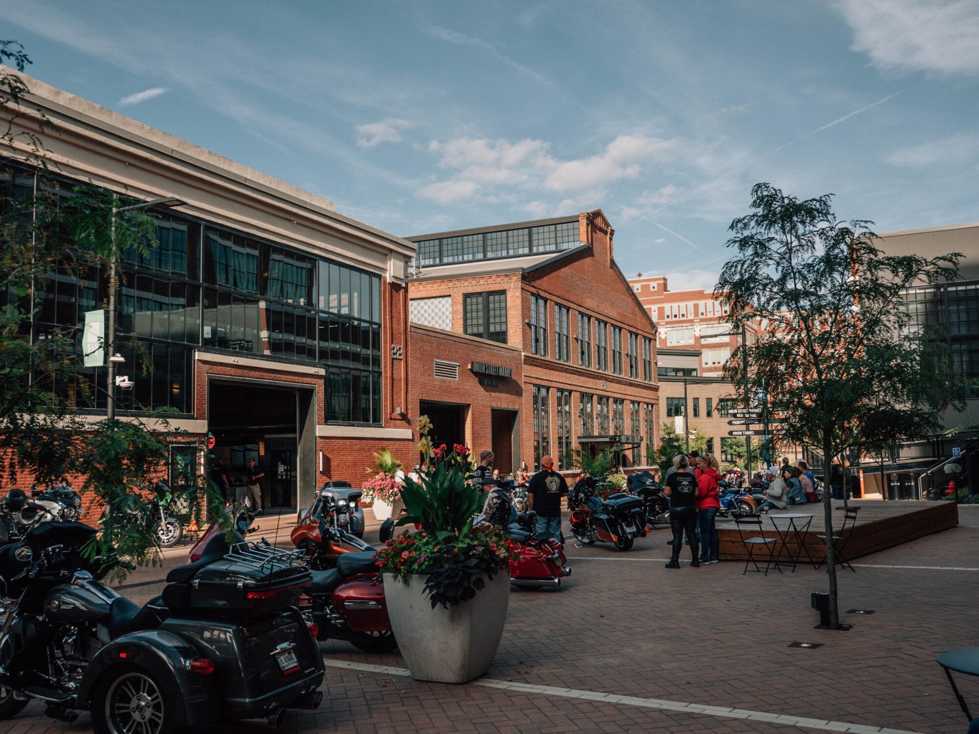 Urban plaza with brick buildings, motorcycles parked along the street, and large flower planters; people gather near the storefronts under a blue sky.